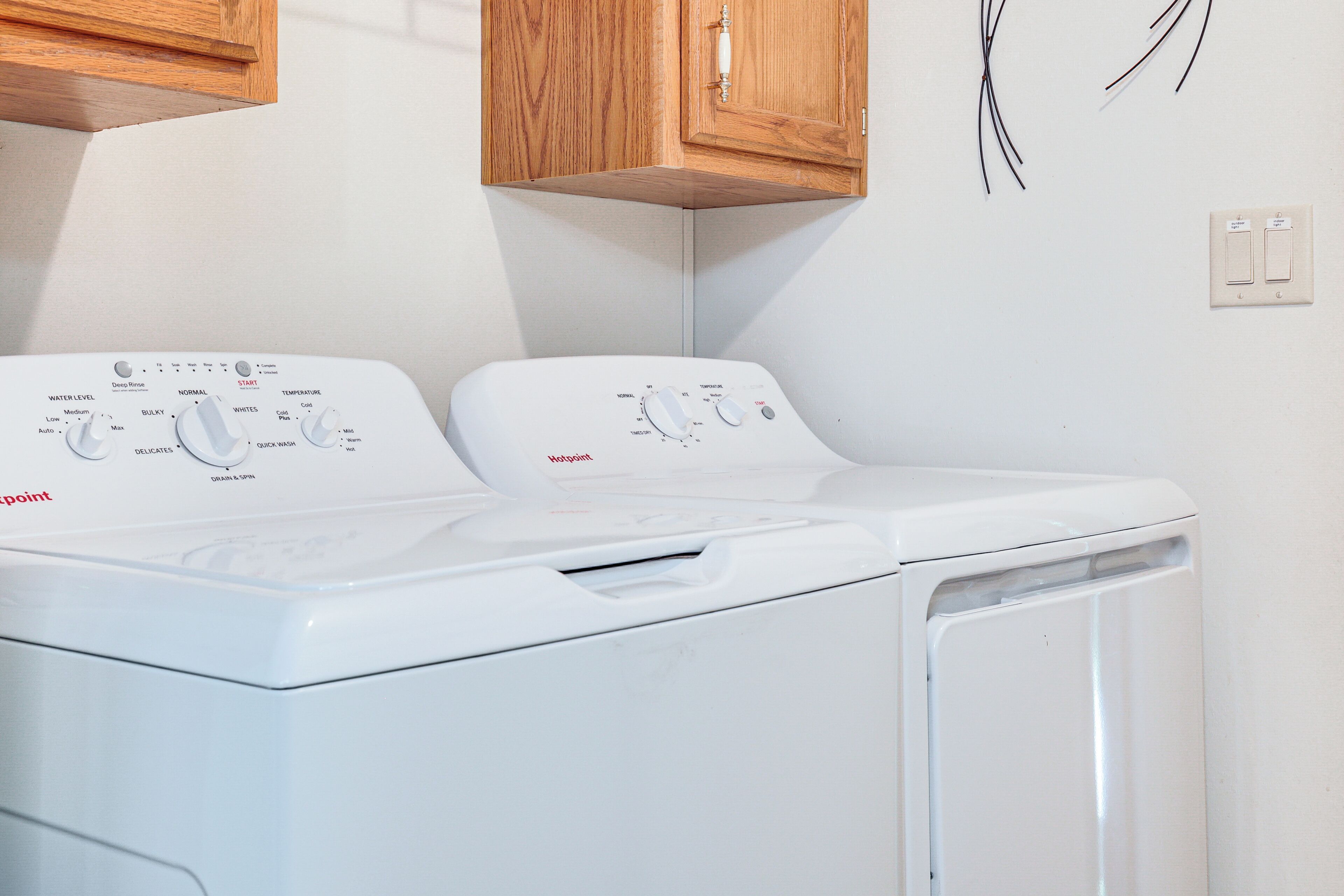A front-loading white washer and dryer unit with control panels, a wooden cabinet above, and a power outlet on the wall.