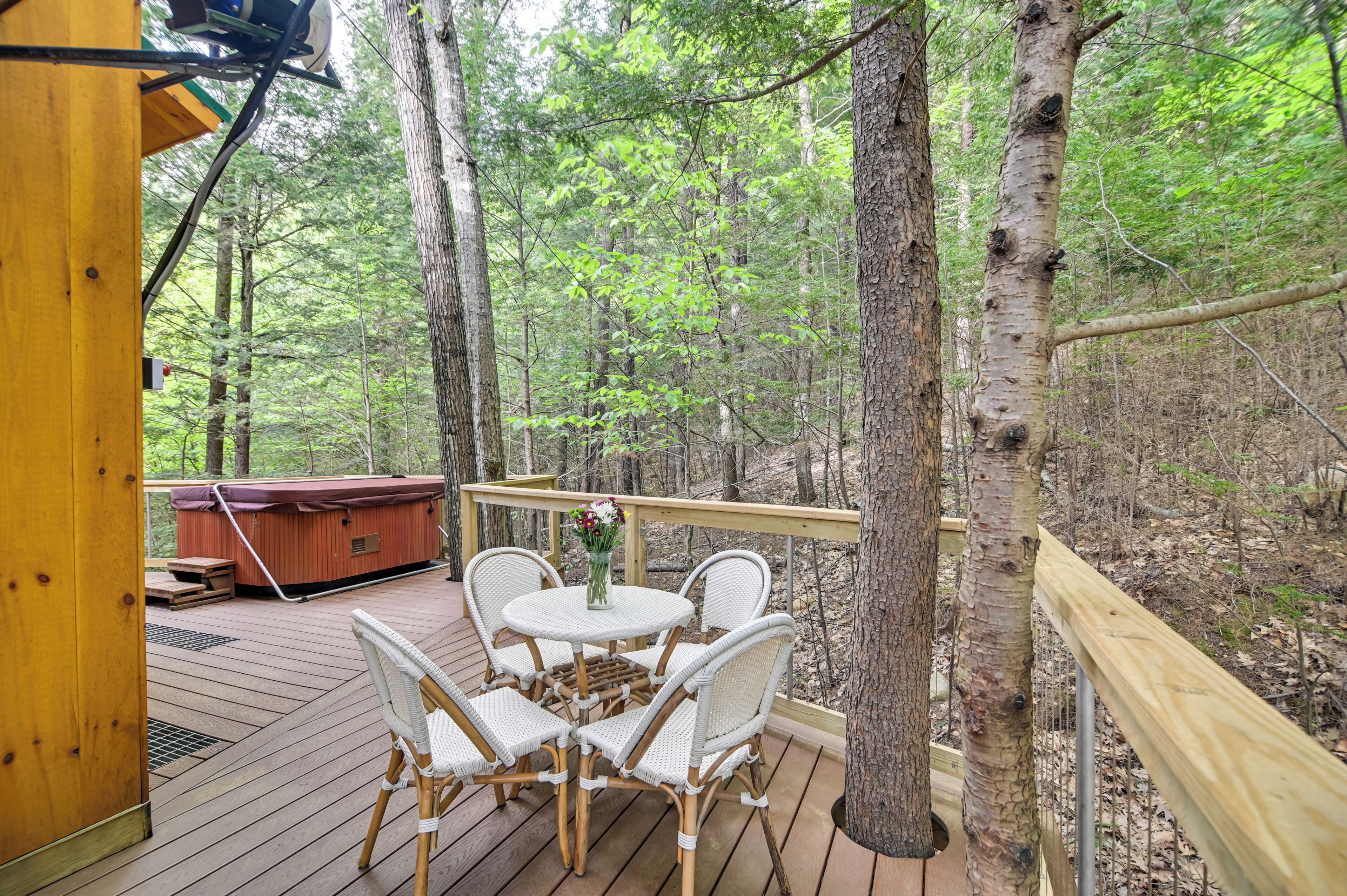 A wooden deck with a hot tub, white wicker chairs, and a table with a vase of flowers.