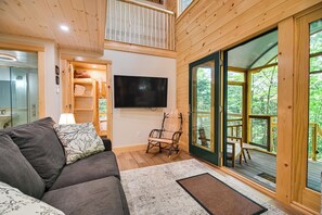 Ein gemütliches Wohnzimmer mit einer Couch, einem Fernseher und einem Holzstuhl. Ein großes Fenster bietet Blick ins Freie.