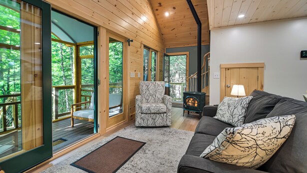 A cozy cabin interior with a wooden ceiling, a sofa with patterned cushions, a chair, and a fireplace. - Tree House w/ Hot Tub: Near Sunday River Resort! (Rumford)