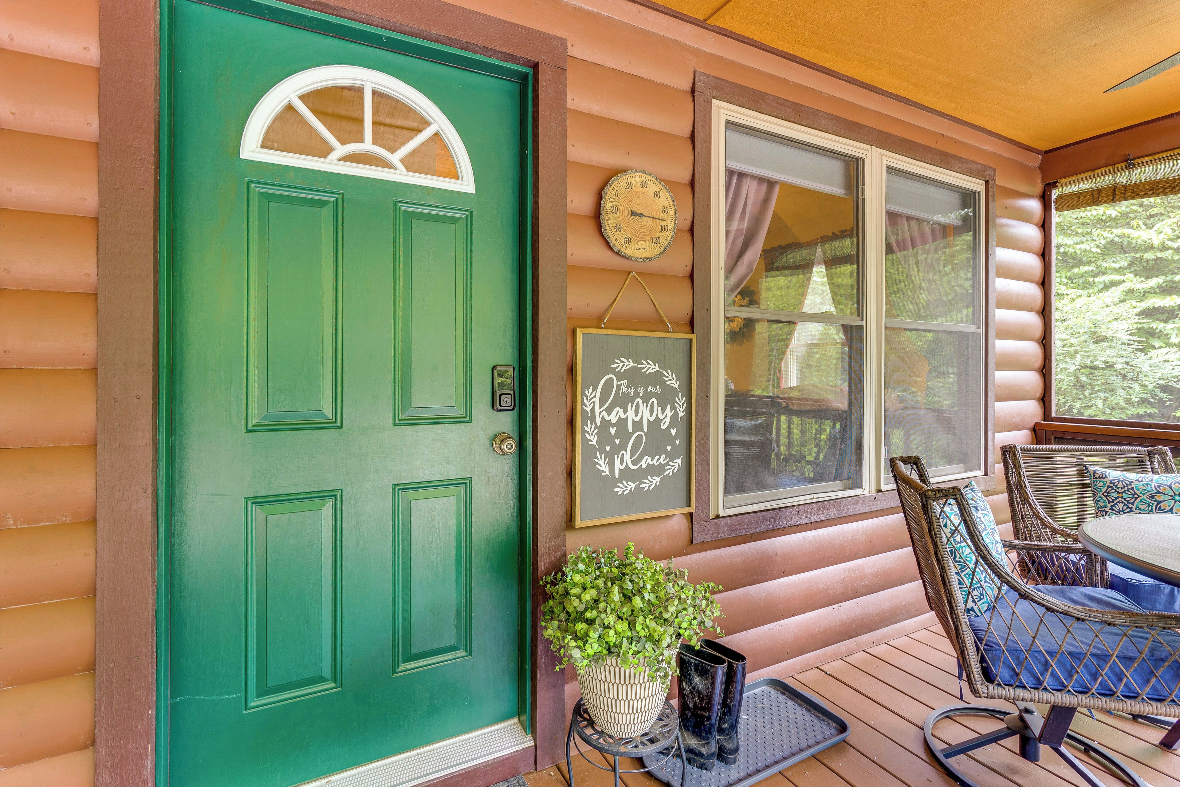A green door with a white arch window, a wall clock, a 'Happy Place' sign, wicker furniture, and a potted plant on a porch.