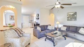 A modern living room with a grey sofa, a patterned armchair, and a coffee table. There is a ceiling fan, a framed picture on the wall, and a kitchen in the background. - 5 Mi to CO River: Home w/ Pool in Fort Mohave! (Fort Mohave)