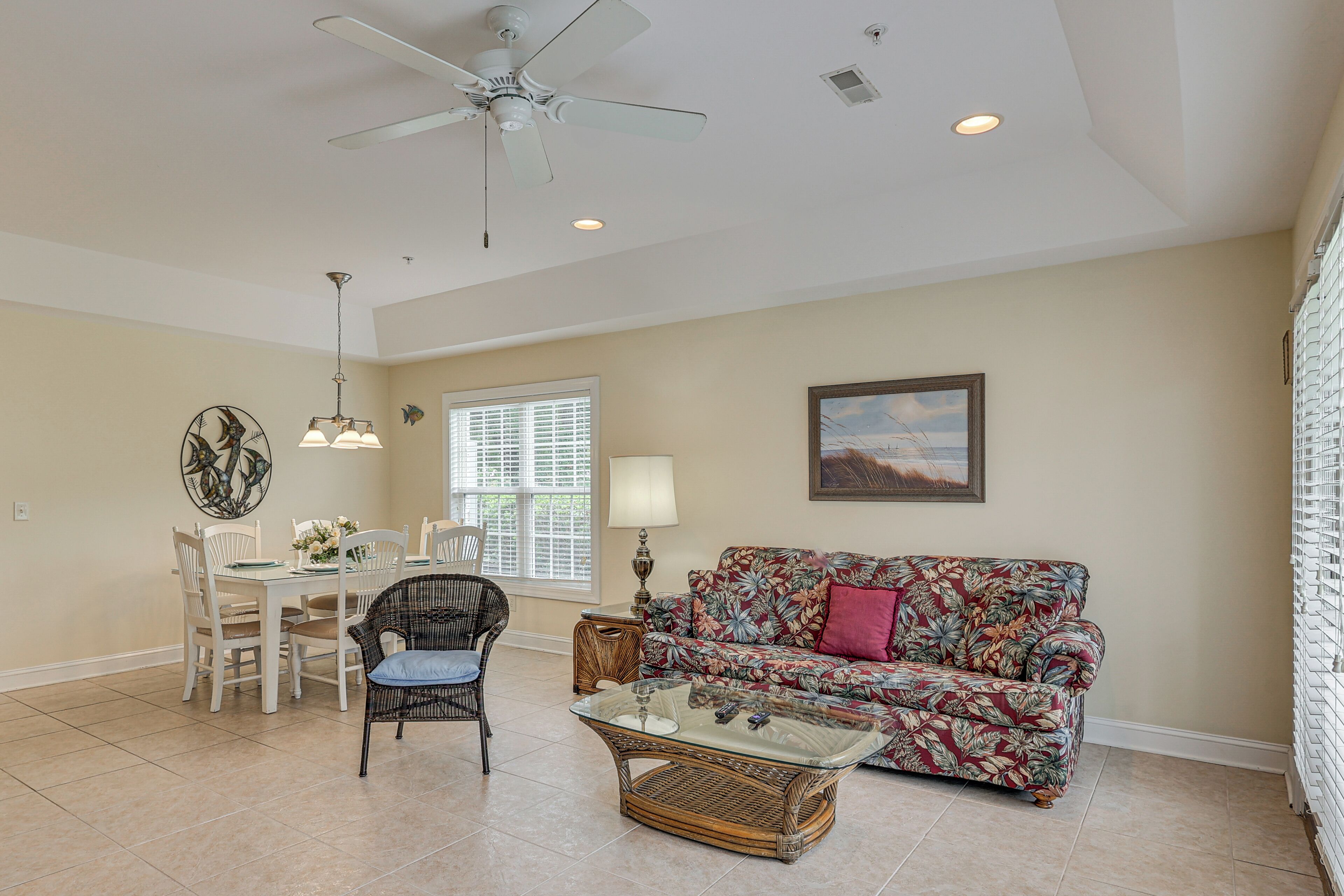 A spacious living room with a ceiling fan, a dining area with a table and chairs, a floral sofa, and a glass coffee table.