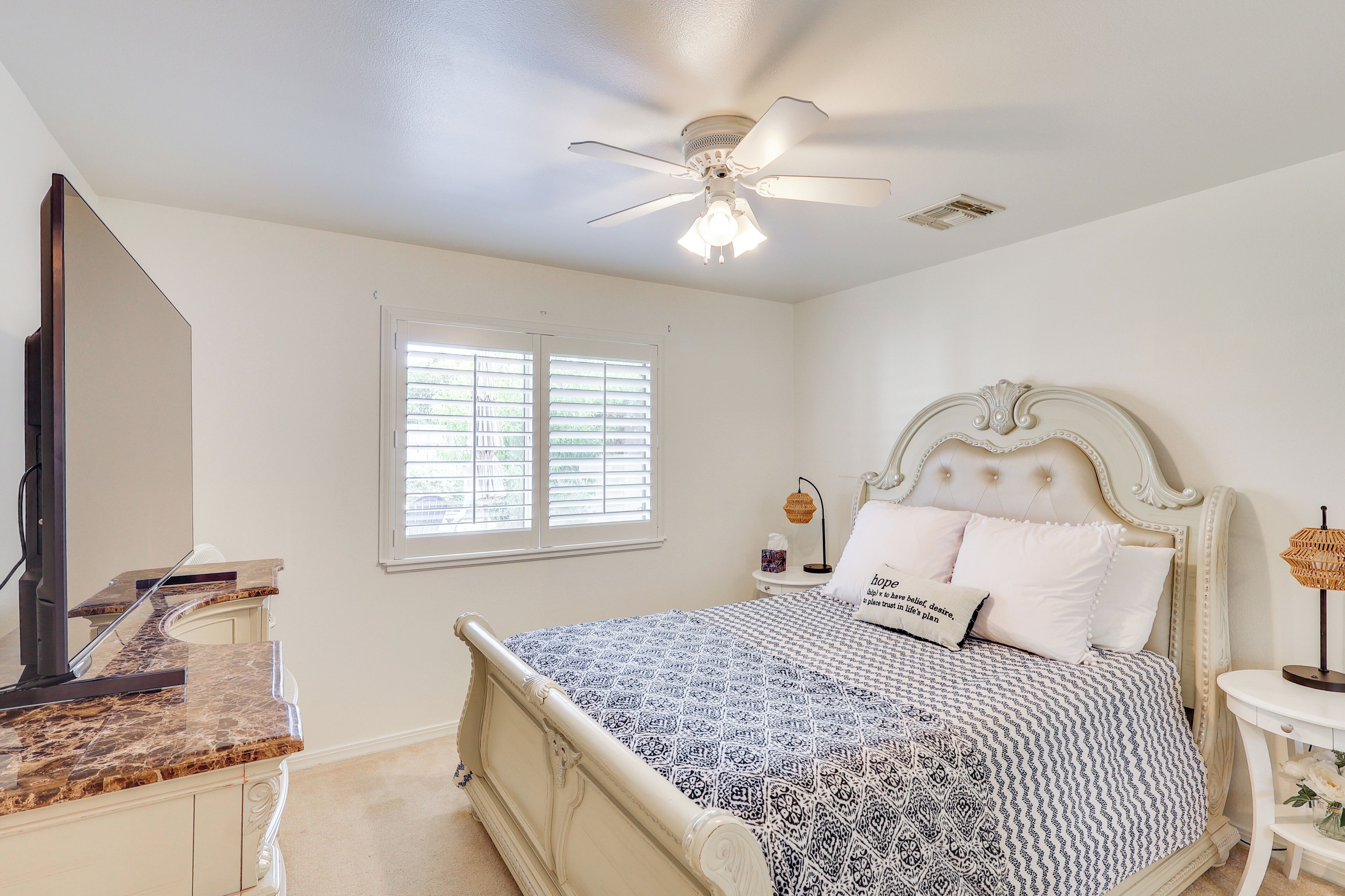 Une chambre à coucher comprenant un lit, un ventilateur de plafond, une fenêtre avec des stores, une table de chevet avec une lampe et une commode avec un miroir.