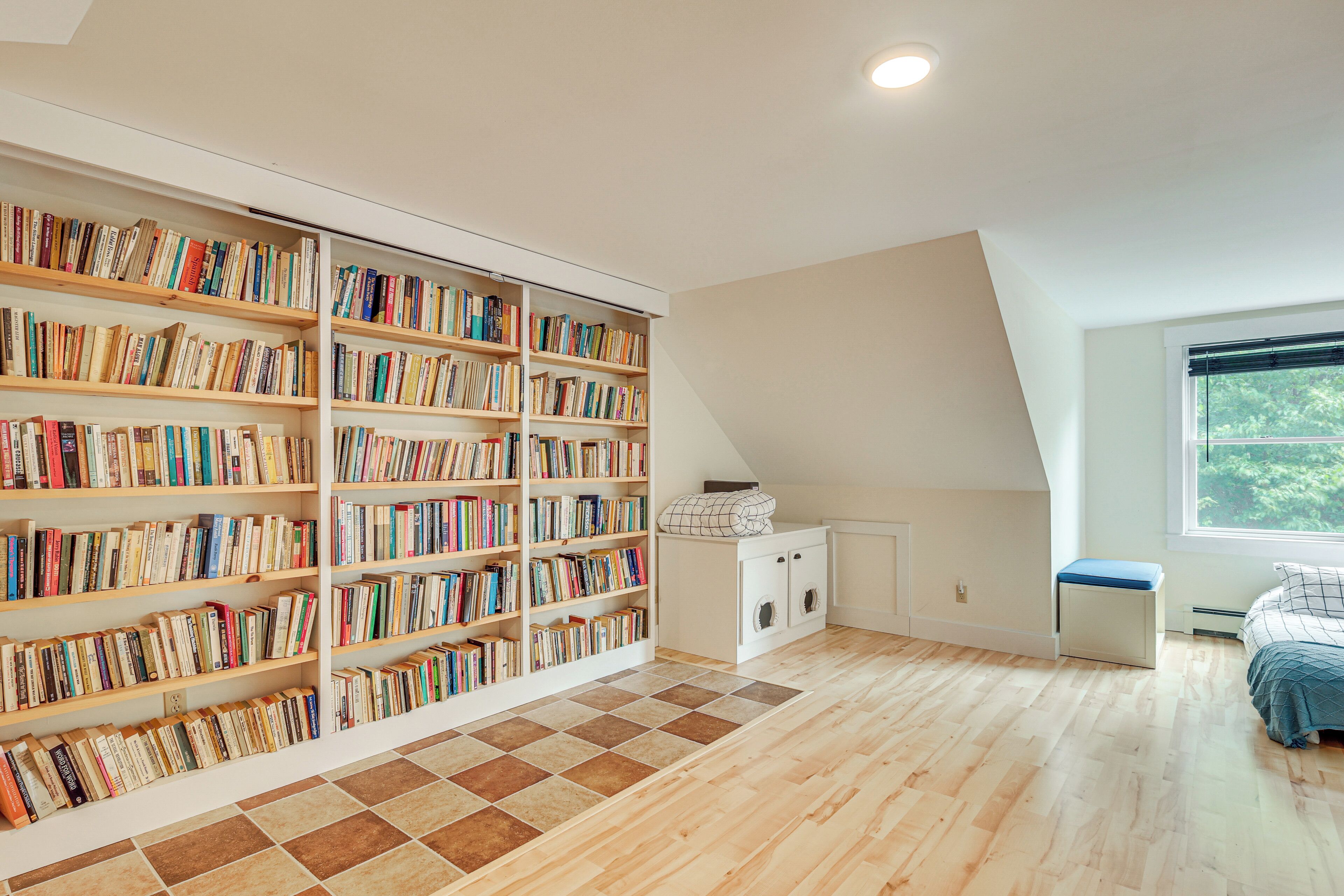 A spacious room with a large bookshelf filled with books, a wooden floor, a window with a view, and a bed with a blue blanket.