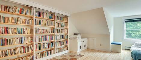 A spacious room with a large bookshelf filled with books, a wooden floor, a window with a view, and a bed with a blue blanket.