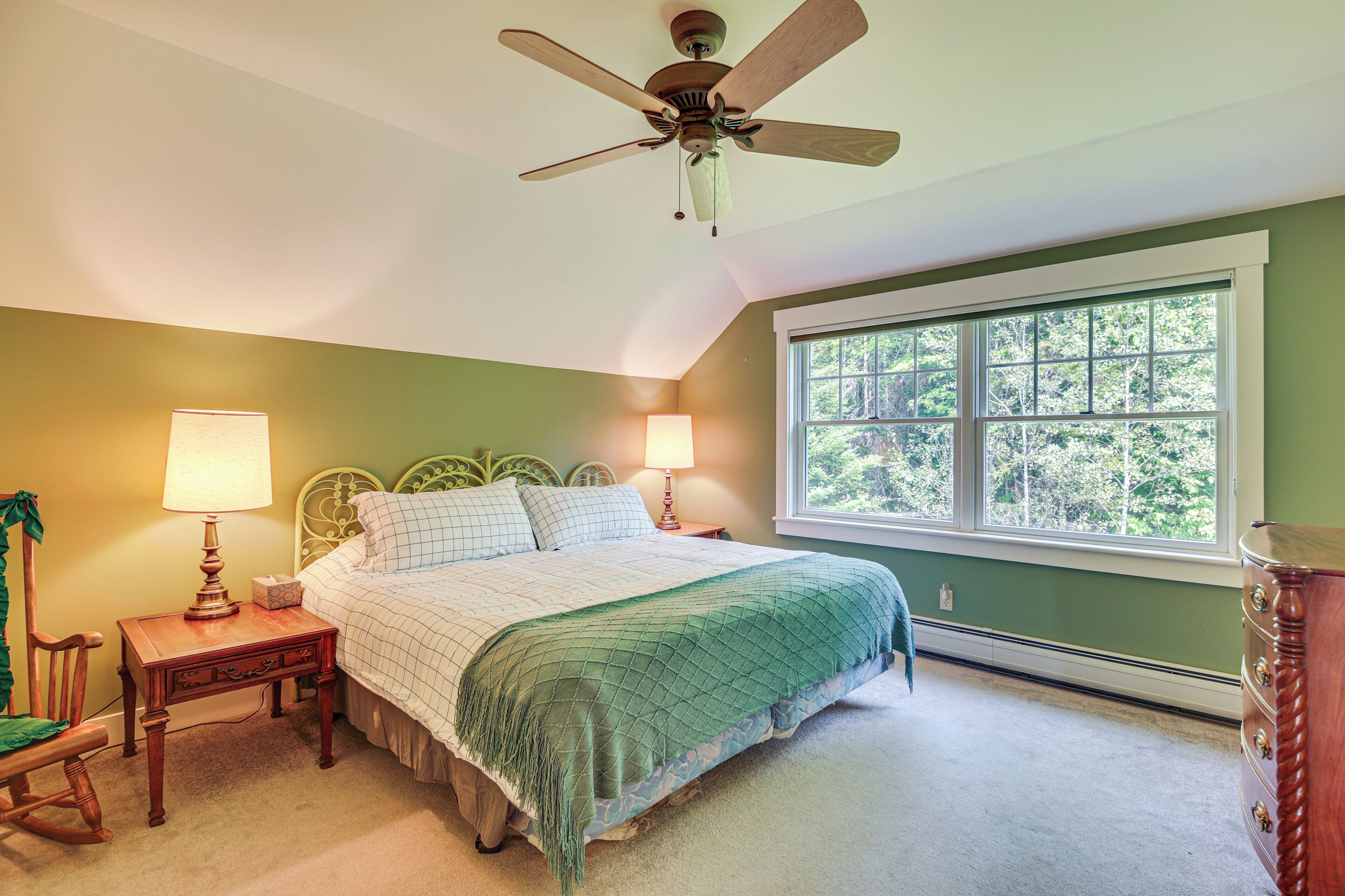 A bedroom with a bed, two bedside lamps, a wooden nightstand, a rocking chair, a dresser, and a window with a view of trees.