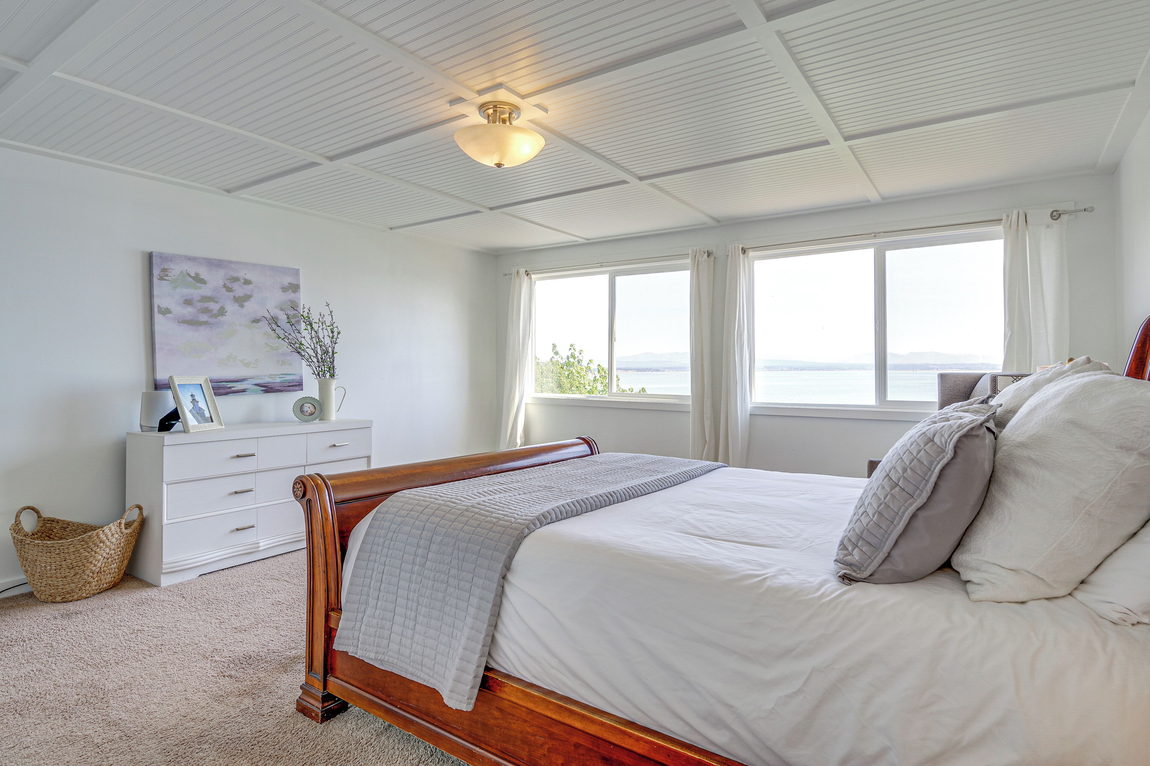 A bedroom with a wooden bed, white bedding, a white dresser, a wall-mounted artwork, and a view of the sea through large windows.