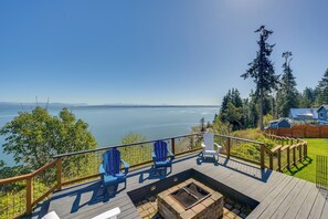 Eine Terrasse mit Stühlen, Blick auf einen See, eine Feuerstelle sowie ein Haus und ein Wald.
