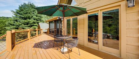 A wooden deck with a table and chairs, a green umbrella, and a view of a forested area.