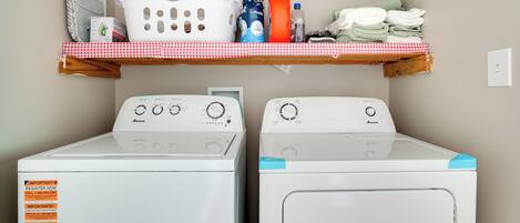 Un espace buanderie comprenant deux machines à laver, une étagère avec des produits de lessive et un tissu à carreaux rouges et blancs.