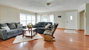 Un salon spacieux doté d’un parquet, d’un canapé gris, d’un fauteuil à carreaux bleus et d’un tapis blanc.
