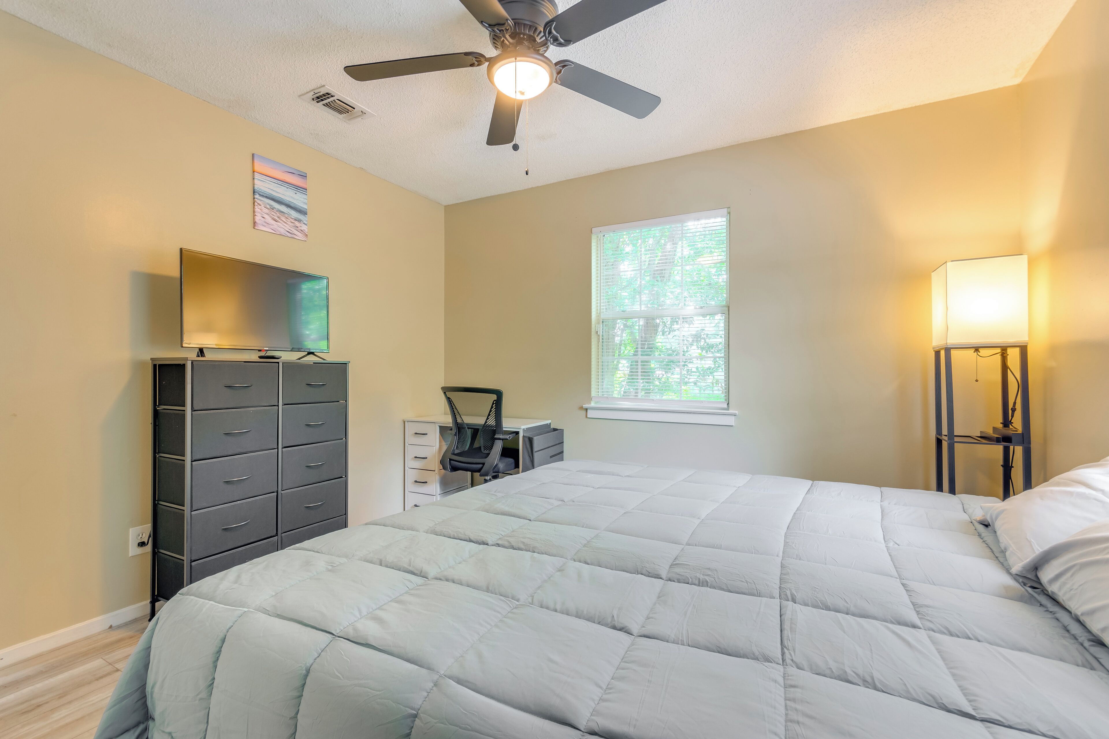 A bedroom with a bed, a dresser, a desk, a chair, a ceiling fan, and a window.