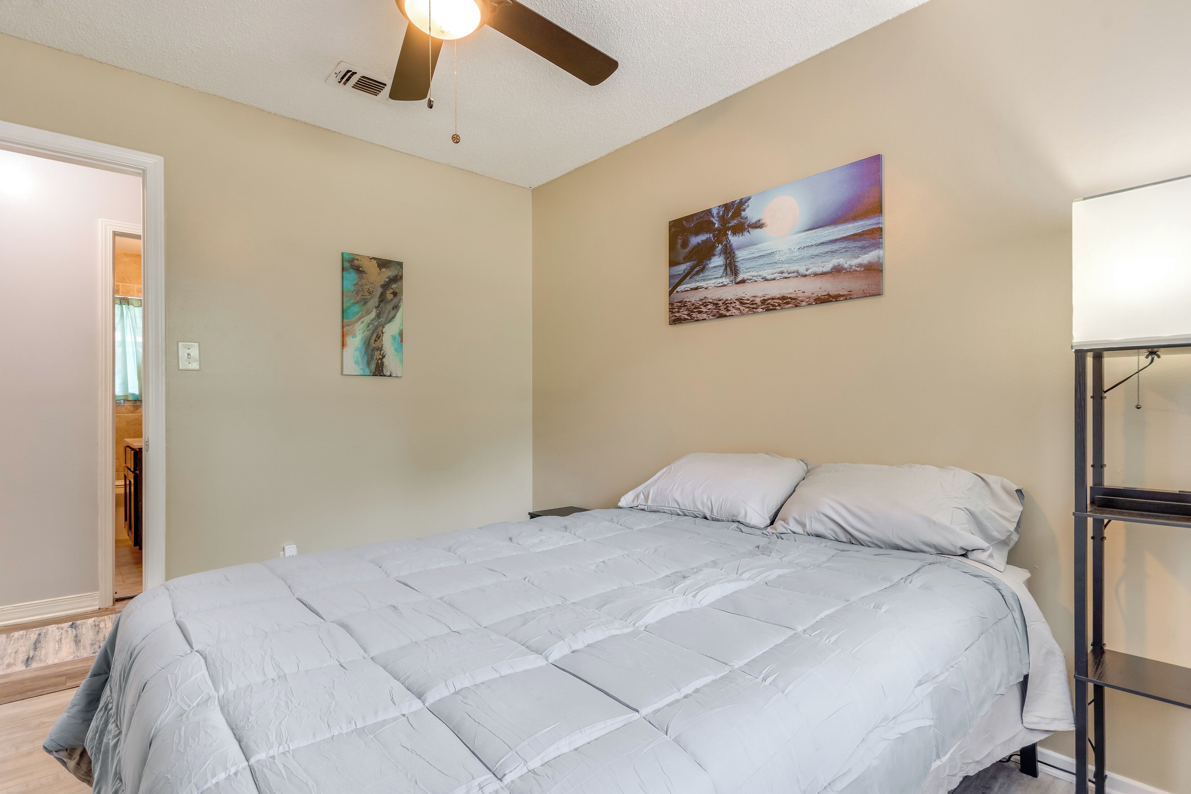 A bedroom with a bed, a ceiling fan, a painting of a beach scene, and a white shelf unit.