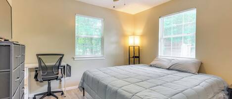 A bedroom with a bed, a desk with a chair, a ceiling fan, and two windows.