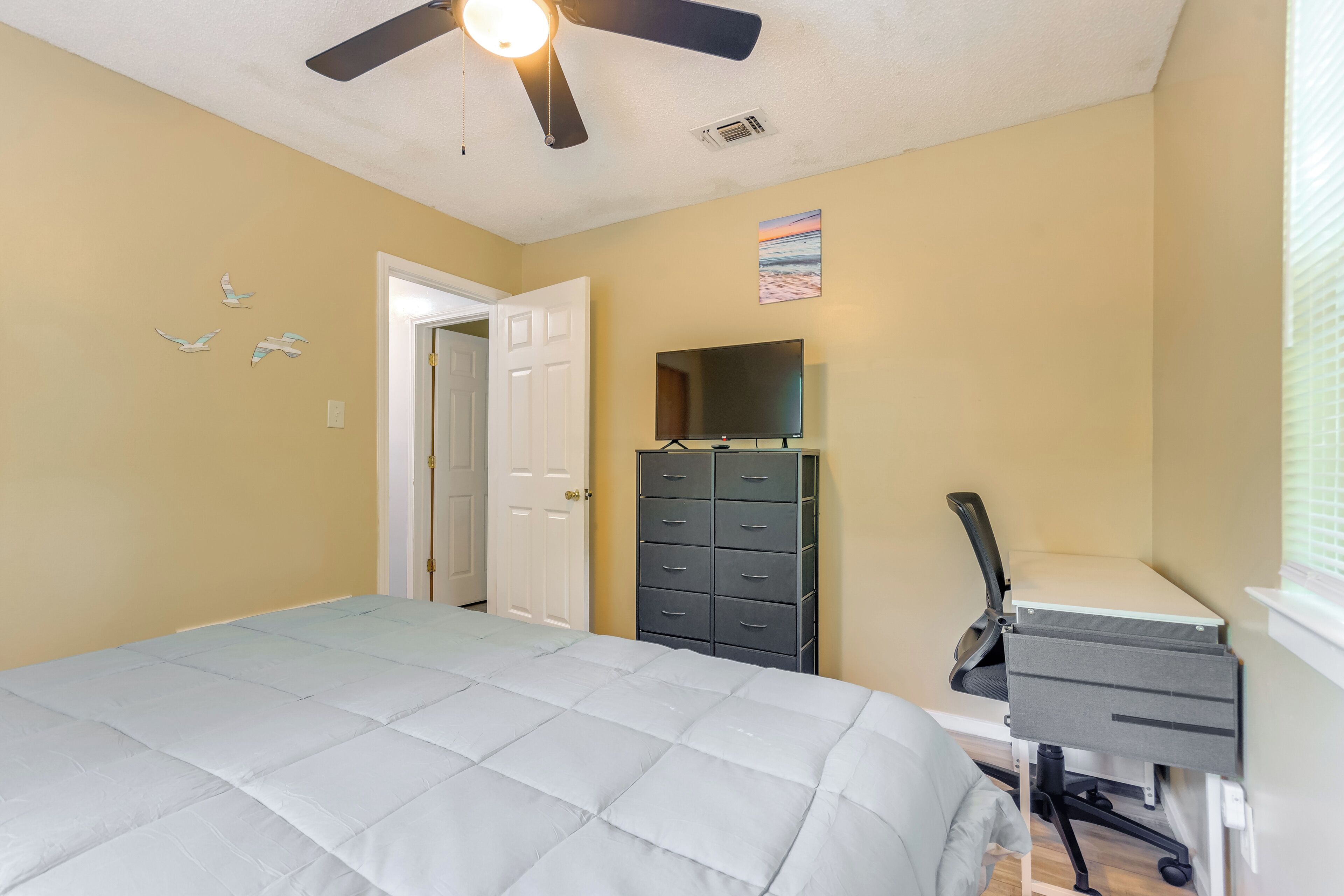 A bedroom with a bed, a desk with a chair, a dresser, a ceiling fan, and a door.