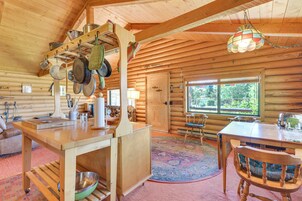 A log cabin interior with a wooden dining table, chairs, a kitchen area with hanging pots, a large window, and a colorful rug.
