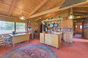 A rustic cabin interior with a wooden dining table, a kitchen area with hanging pots, and a large rug.