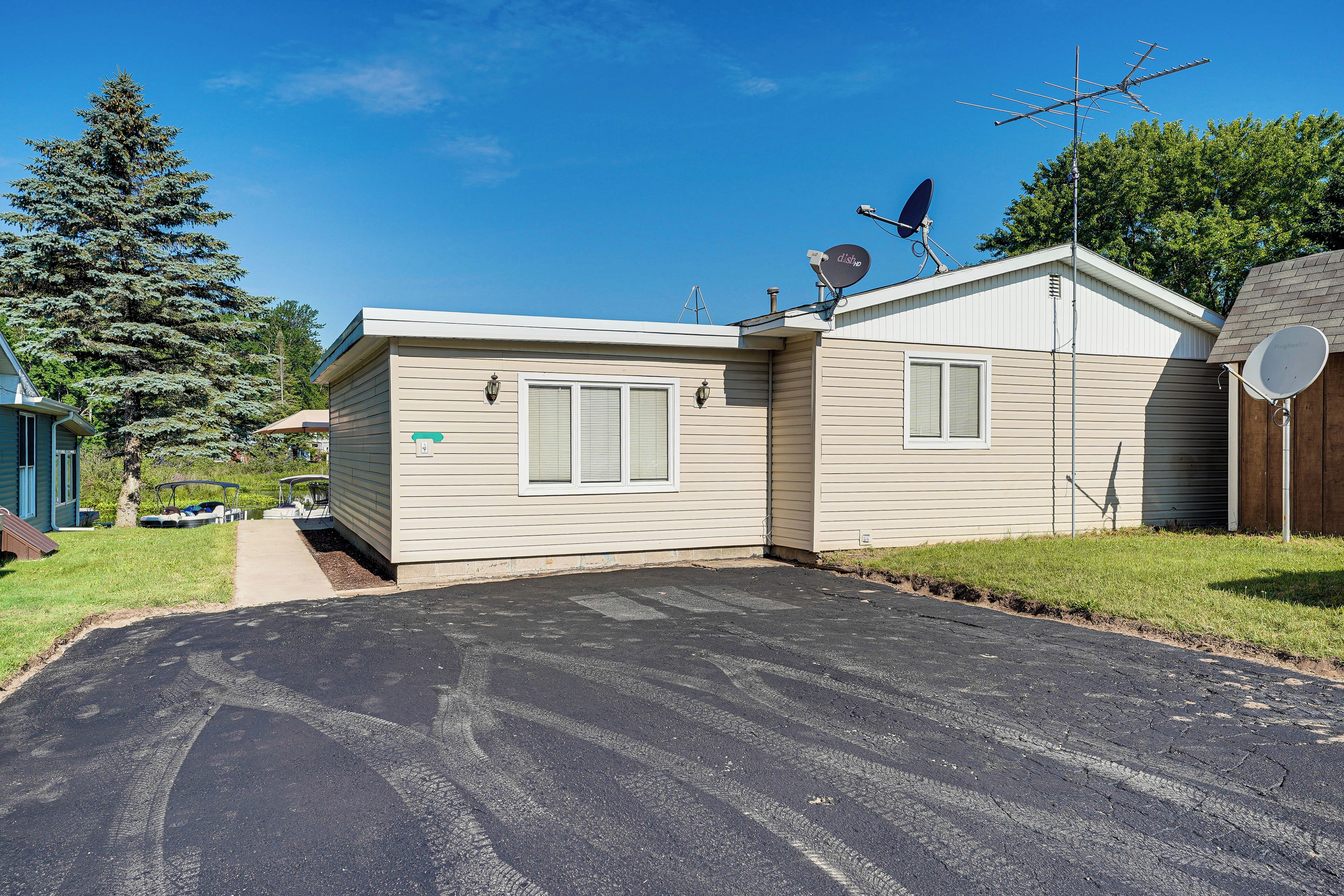 A single-story mobile home with a driveway, satellite dishes, and a large tree in the background.