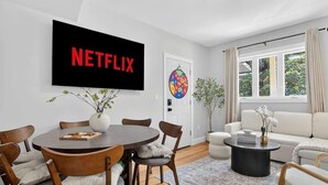Ein modernes Wohnzimmer mit Essbereich, einem Wand-Fernseher, einer Dartscheibe und einem großen Fenster mit Vorhängen.