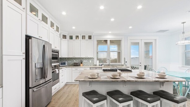 A modern kitchen with white cabinetry, a stainless steel refrigerator, a central island with stools, and a view to the outside. - Heated Pool Elevator Game Room Ocean Decks (St. Augustine)
