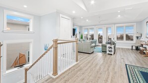 A spacious living area with a wooden floor, a staircase with a white railing, and a view of the outside through large windows. - Heated Pool Elevator Game Room Ocean Decks (St. Augustine)