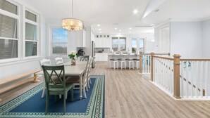 A spacious kitchen with white cabinets, a dining area with a blue rug, and a staircase with wooden railings. - Heated Pool Elevator Game Room Ocean Decks (St. Augustine)