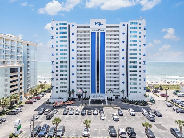 A multi-story hotel with a central entrance, parking lot, and a view of the beach.