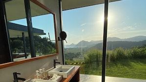 Un baño moderno con dos lavamanos, un espejo grande y vistas a un paisaje verde a través de amplios ventanales.