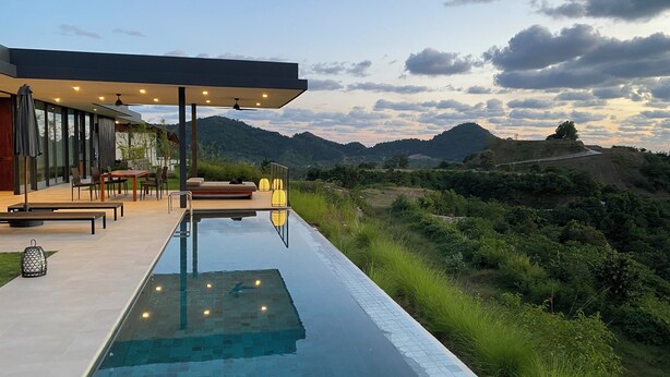 Un área de piscina moderna al aire libre con una zona de estar cubierta, mobiliario para comer y vistas del paisaje circundante.