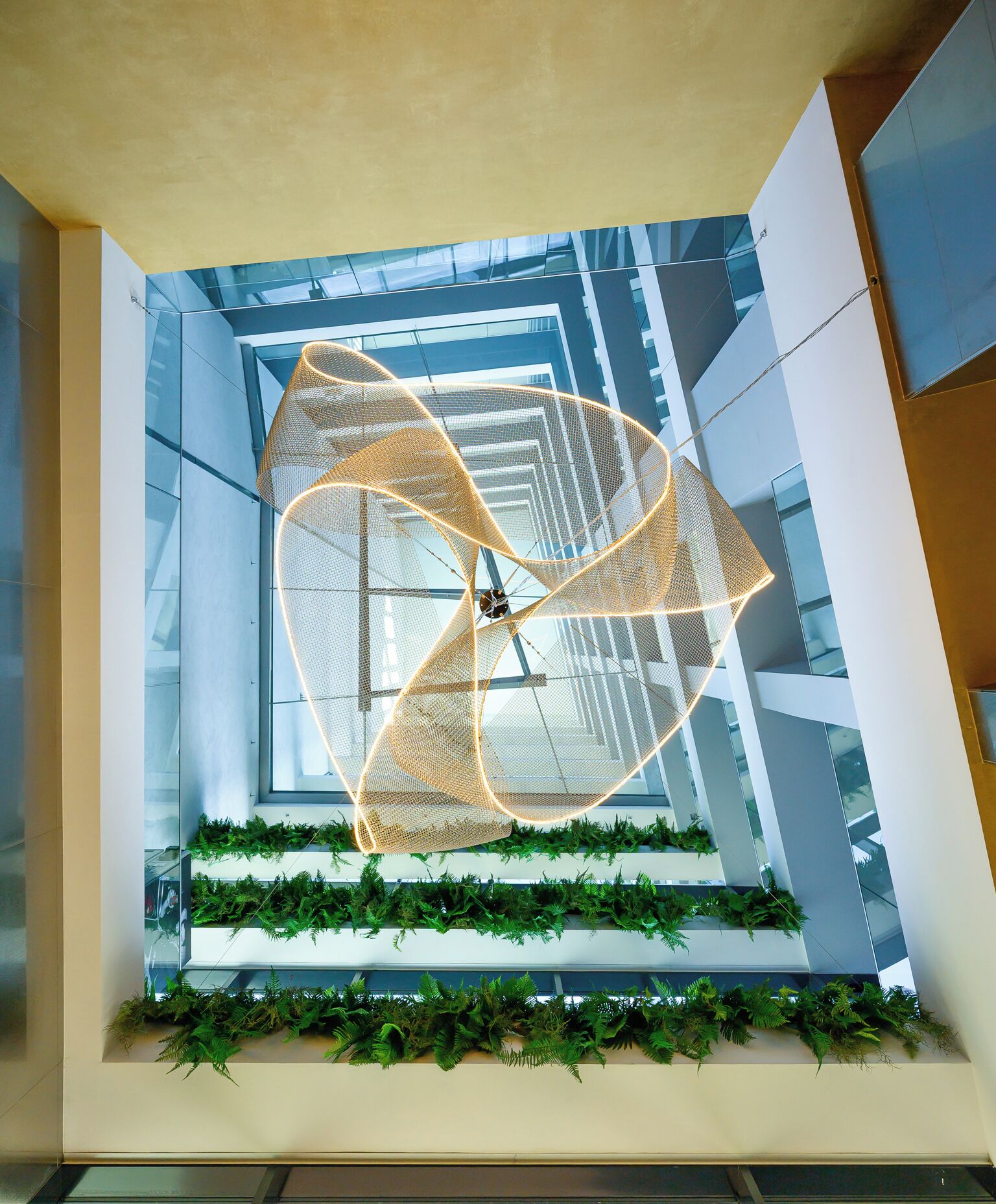 A modern atrium with a geometric light fixture, glass walls, and green plant installations.