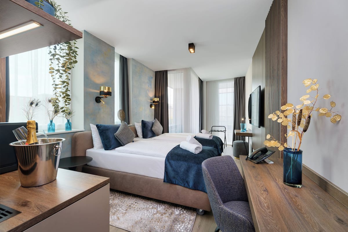 A modern hotel room with a bed, bedside table, and a desk with a chair. There is a large window, a plant, and a vase with flowers.