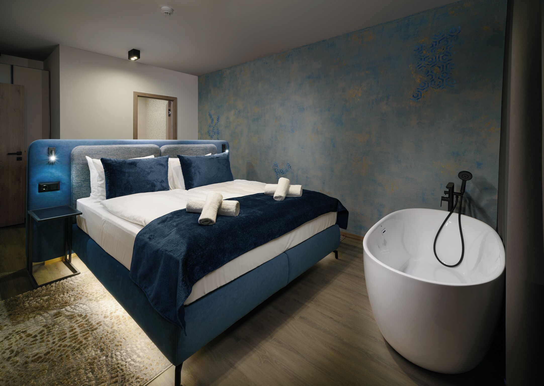 Quarto moderno com cama grande, mesa de cabeceira, banheira embutida e parede texturizada.