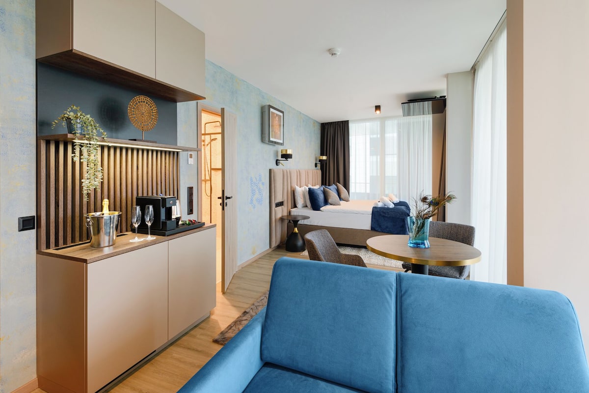 A modern hotel room with a kitchenette, a bed with a view, a blue sofa, and a dining area.
