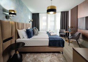 Moderni hotellihuone, jossa on suuri sänky, yöpöydät, televisio sekä oleskelualue, jossa on tuoli ja pöytä.
