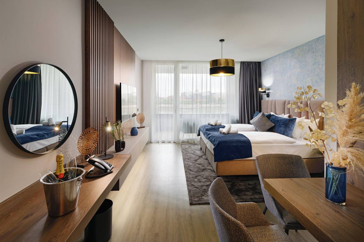A modern hotel room with a large bed, a desk with a telephone, a mirror, and a dining area with a table and chairs.