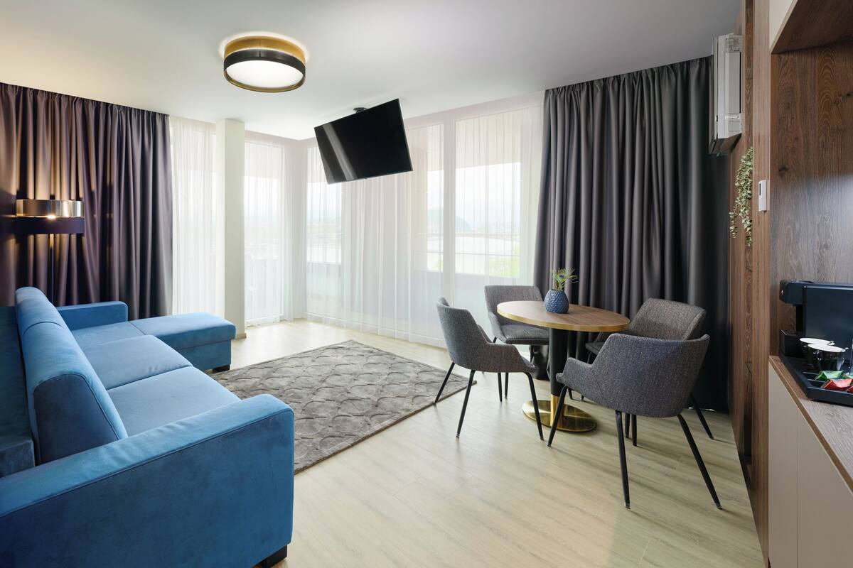 A modern living room with a blue sofa set, a small round table with chairs, a flat-screen TV mounted on the wall, and large windows with sheer curtains.