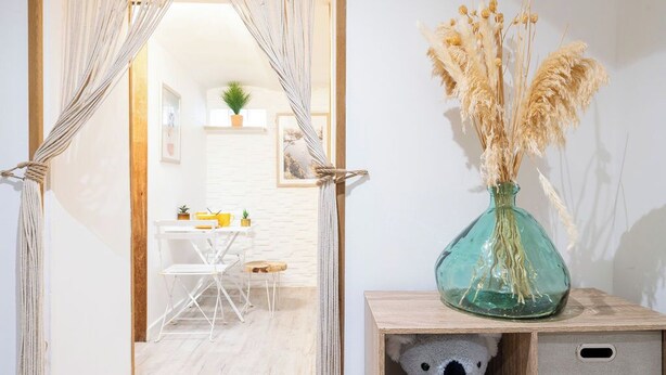 Une porte surmontée d’une tenture en macramé, une petite table avec des chaises et un vase contenant des plantes séchées, posé sur une table d’appoint en bois.