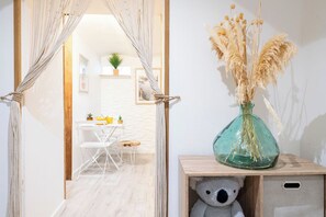 Une porte surmontée d’une tenture en macramé, une petite table avec des chaises et un vase contenant des plantes séchées, posé sur une table d’appoint en bois.