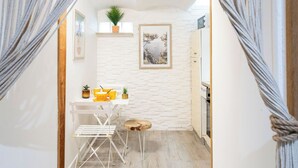 Un couloir étroit avec un mur en briques blanches, une petite table sur laquelle sont posés des objets jaunes, une plante et un tableau encadré.