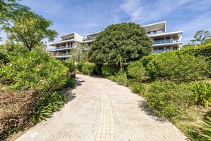 A pathway leading to a modern building with multiple balconies, surrounded by lush greenery.