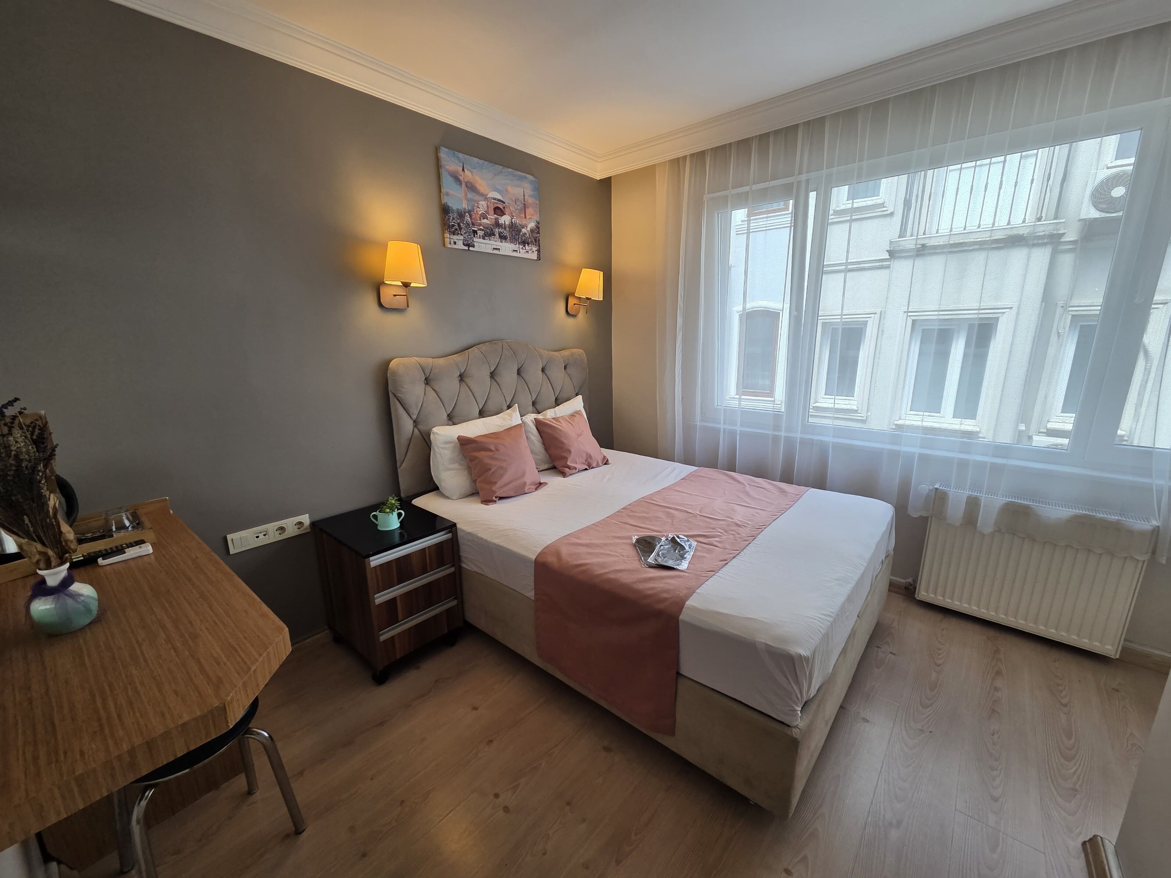 A hotel room with a bed, bedside lamps, a painting on the wall, a window with a view of buildings, and a radiator.