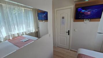 Habitación de hotel con cama, televisor montado en la pared, puerta con un letrero y ventana con cortinas translúcidas.
