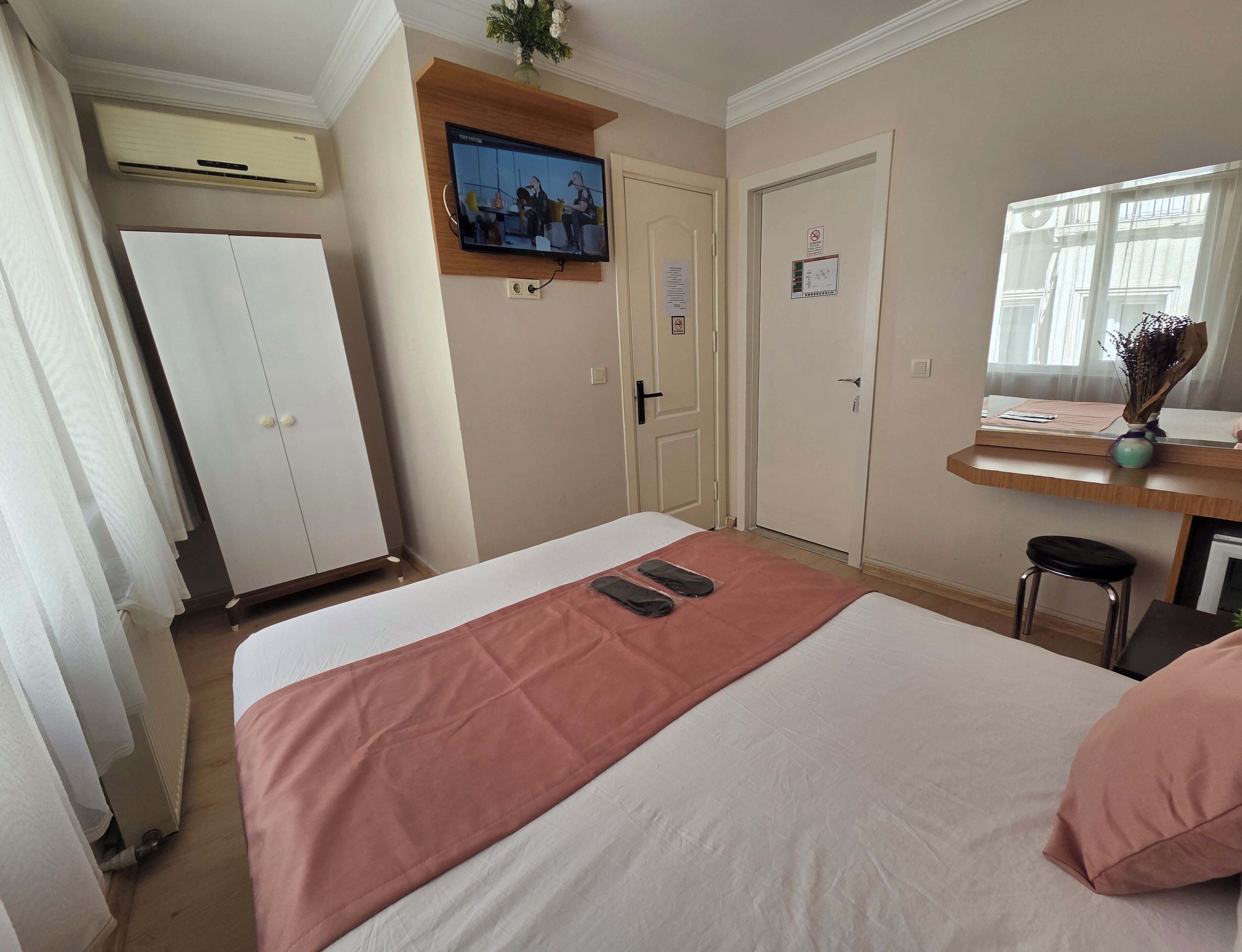 A hotel room with a bed, a TV mounted on the wall, a desk with a vase, and a window with curtains.