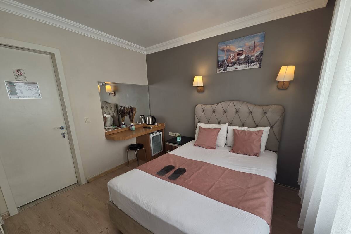 A hotel room with a bed, bedside lamps, a small desk, a chair, a nightstand, a painting, and a door.