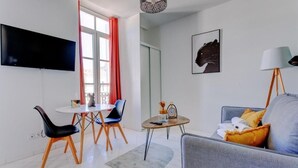 A modern living room with a flat-screen TV, a dining area with chairs, a sofa with cushions, a lamp, and a framed artwork on the wall. - La Panthère studio design central et calme (Cannes)