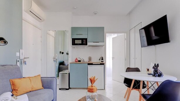 A modern living room with a sofa, a coffee table, and a dining area with chairs and a table. There is a kitchen in the background with a microwave and cabinets. A flat-screen TV is mounted on the wall. - La Panthère studio design central et calme (Cannes)