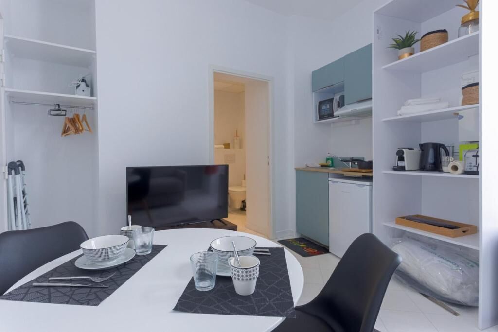 A modern dining area with a white table set for four, a flat-screen TV, and a kitchen area with a microwave and shelves.