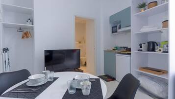 A modern dining area with a white table set for four, a flat-screen TV, and a kitchen area with a microwave and shelves.