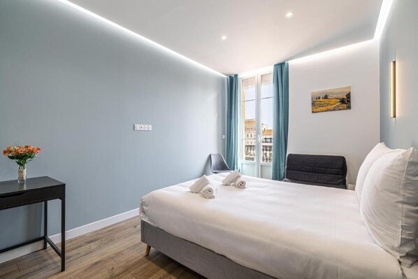 Habitación de hotel moderna con una cama, un escritorio y una silla. Hay una ventana con cortinas, un cuadro en la pared y un florero con flores sobre el escritorio.